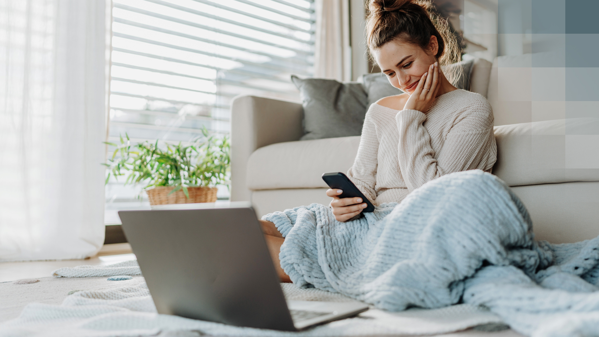 Eine junge Frau sitzt mit einer Decke vor dem Sofa, hält ihr Smartphone und hat ein Laptop vor sich, und informiert sich über die digitale Grundversicherung.